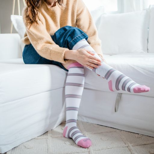 Woman sitting on couch wearing tall white socks with grey and pink stripes, pink top band,heel and toe caps-Motif Medical Gradient Maternity Compression Socks