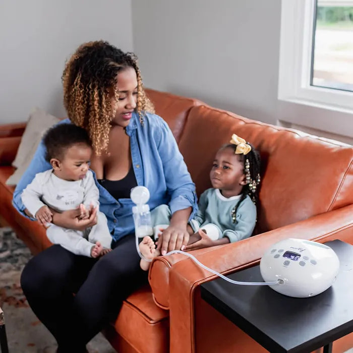 Woman sitting on a brown couch with two children while wearing Hands-Free Pumping Bra and using Motif Medical breast pump