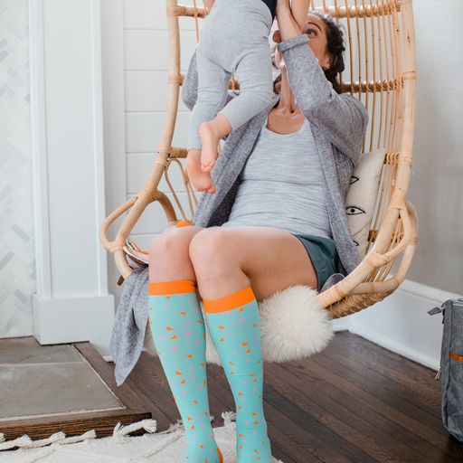 Woman holding child and wearing mint green tall sock with pink and orange triangles and orange band,heel and toe caps -Motif Medical Gradient Maternity Compression Socks